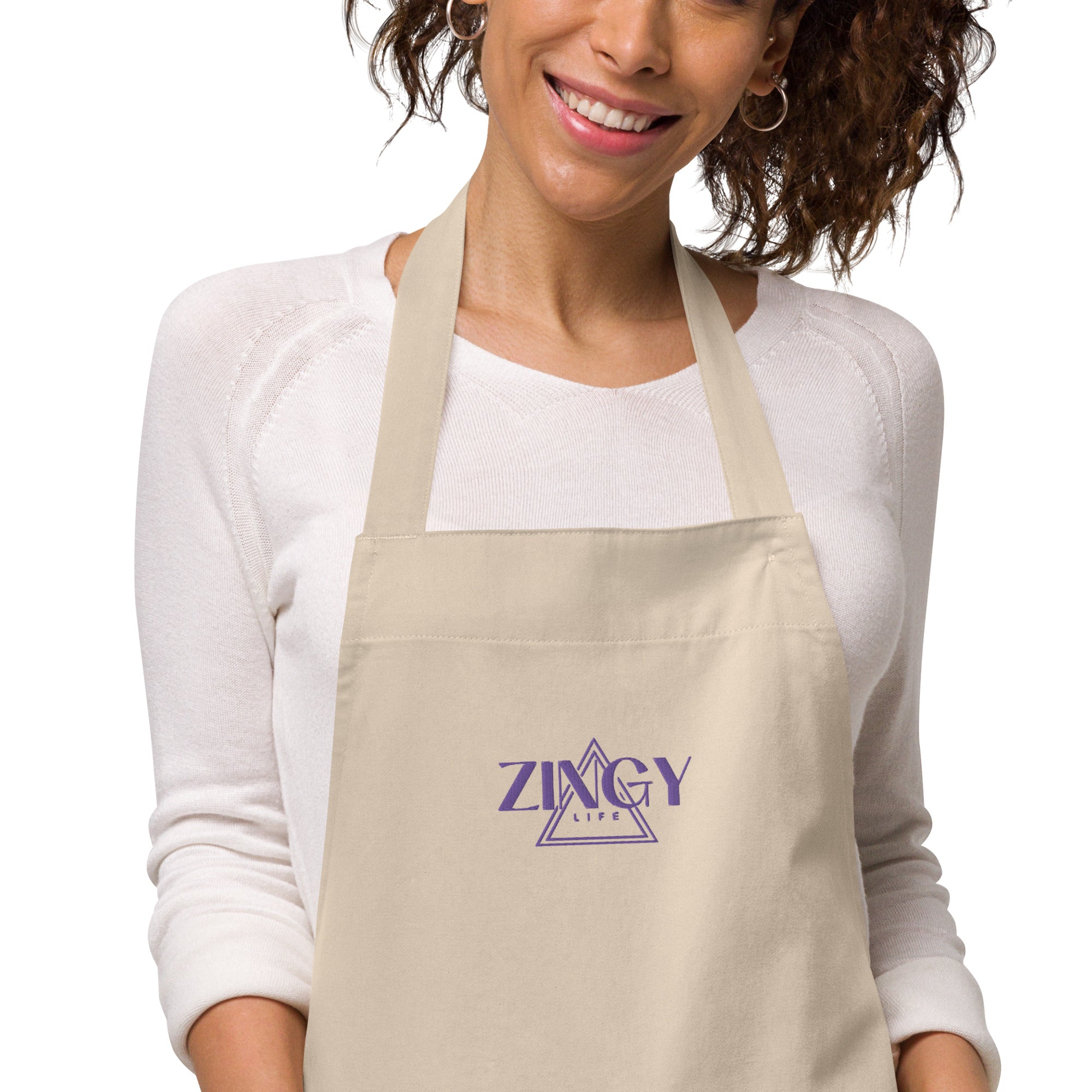 A person wearing a beige organic cotton apron with a large front pocket and adjustable straps. The apron has a brand logo on the front.